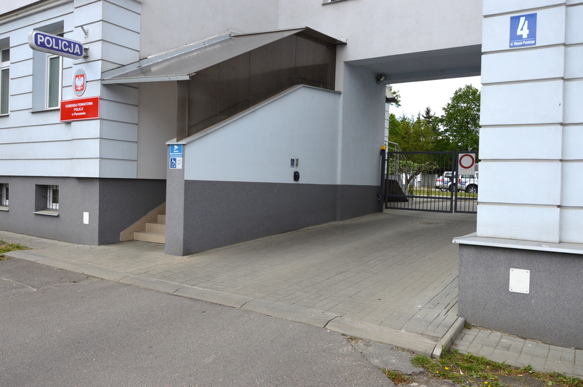 zdjęcie przedstawia wejście głowne do Komendy Powiatowej Policji w Parczewie.Przy schodach wejściowych widnieje tabliczka oznaczona  dla osób z niepełnosprawnością.Obok wejścia znajduje sie brama wjazdowa.Po lewej stronie  na ścianie przy bramie głownej znajduje sie domofon.
