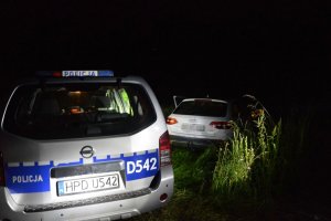 W nocy w terenie niezabudowanym policyjny radiowóz na drugim planie białe audi, którego kierujący zatrzymał się na łące.