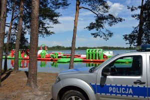 Policyjny radiowóz nad jeziorem w miejscowości Białka. Na drugim planie jezioro i dmuchane elementy do zabaw dla dzieci